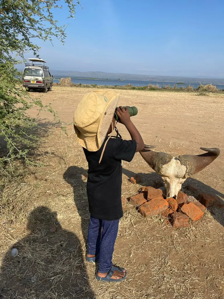 A child in a safari hat looking through binoculars beside a lake, with a safari vehicle nearby