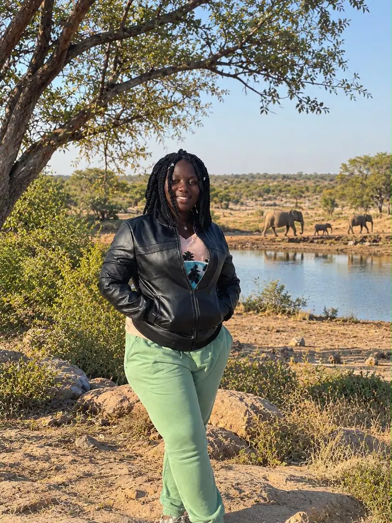 Irene beneath a tree beside a waterhole, with elephants on the opposite bank